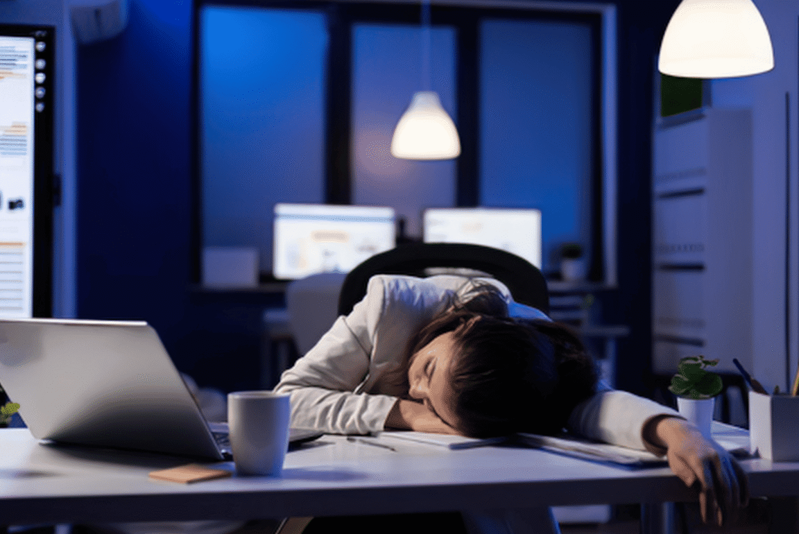 A person asleep with their head resting on a laptop at a desk.